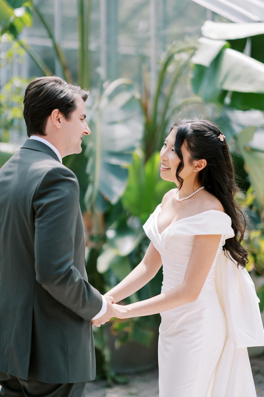 A bride and groom standing and holding hands in a lush garden setting, with greenery and natural light enhancing the romantic atmosphere of their wedding day.