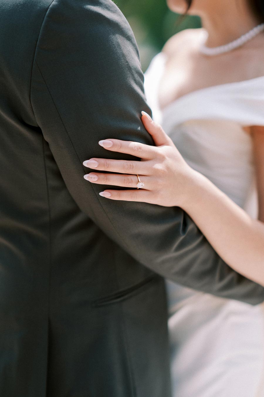 Elegant bride in white wedding dress embraces groom in dark suit, highlighting engagement ring on left hand.