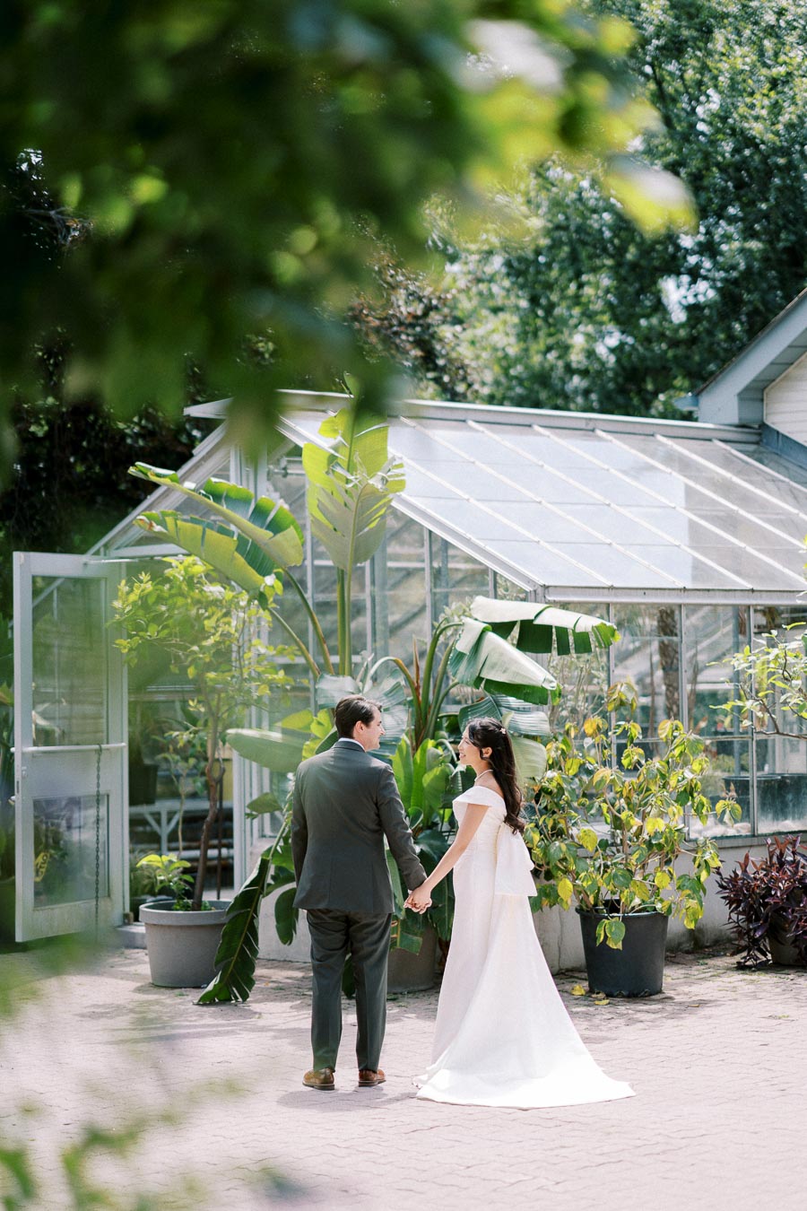 Wedding couple holding hands in front of lush greenhouse with tropical plants, capturing a romantic outdoor ceremony atmosphere.