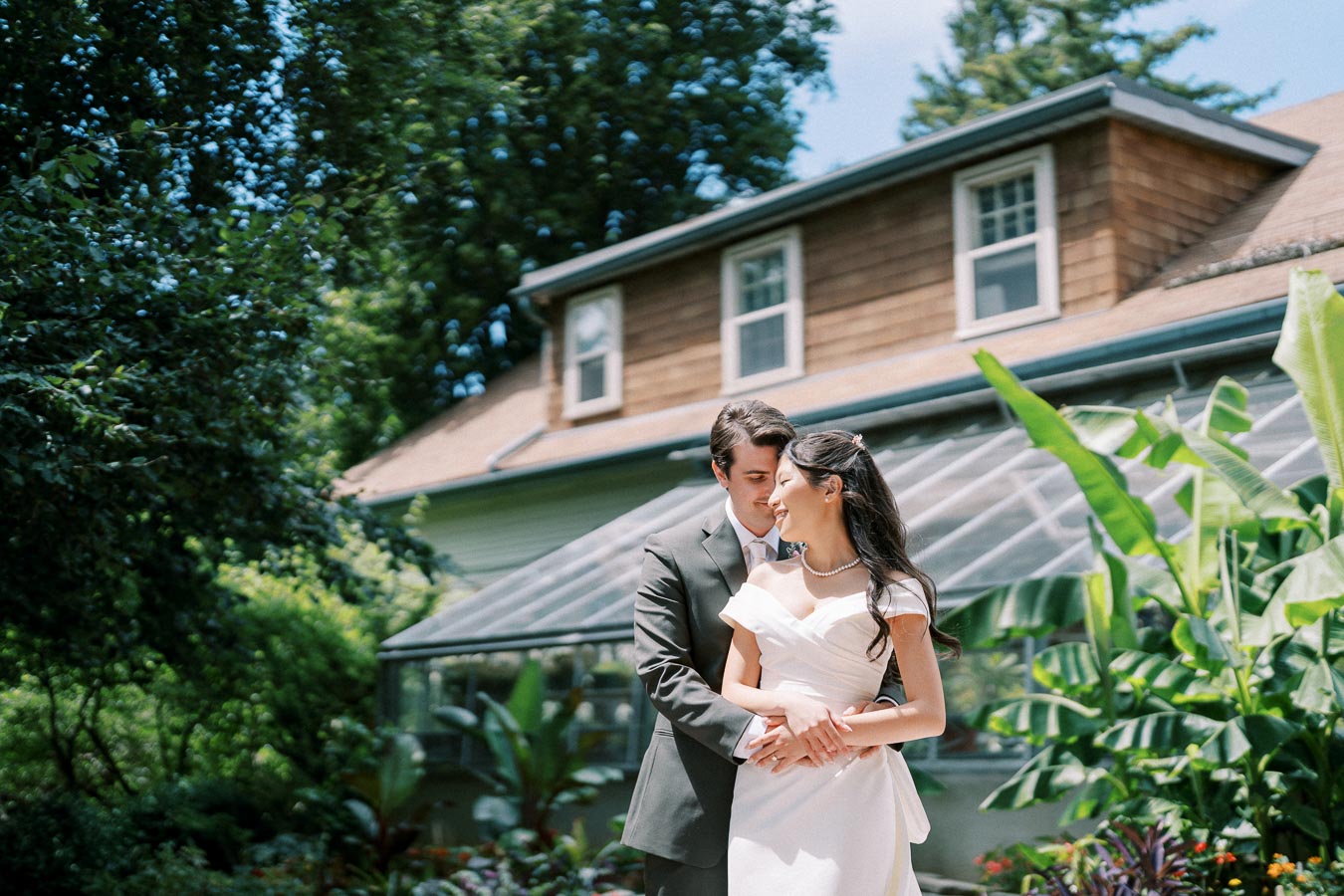 Elegant bride and groom embracing in garden setting by rustic house, surrounded by lush greenery and vibrant plants, under a clear blue sky.