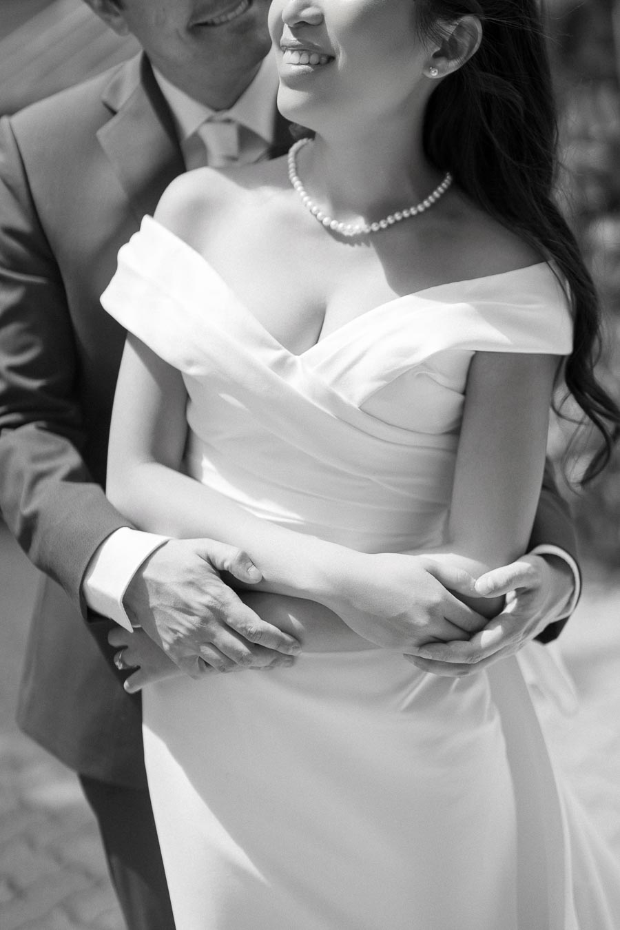 A black and white photograph of a couple embracing on their wedding day, focusing on the bride with a pearl necklace and an elegant off-the-shoulder gown, and the groom in a suit, highlighting their hands intertwined around her waist, symbolizing love and togetherness.