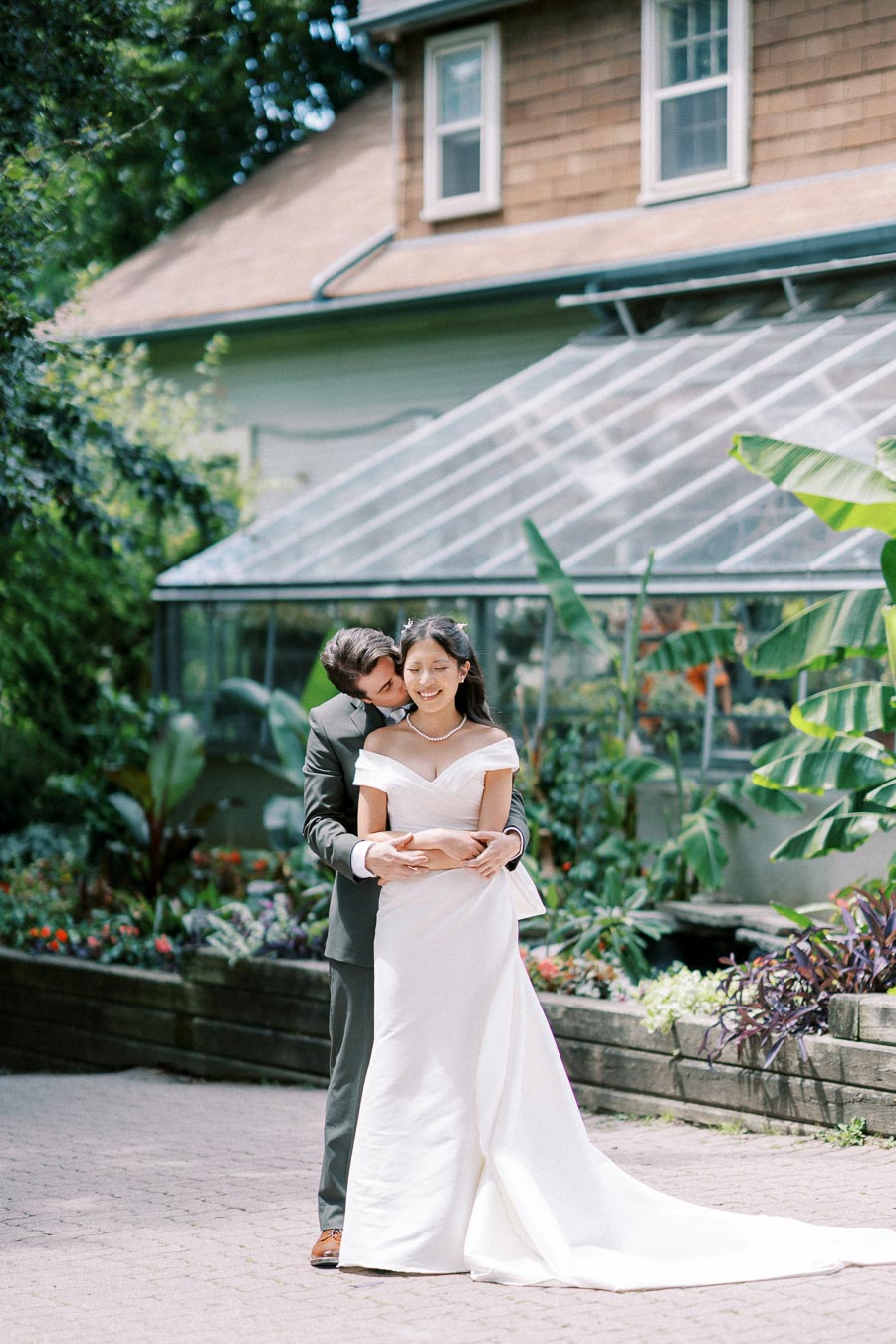 Bride and groom embracing in a garden setting with a greenhouse background, capturing a joyful wedding moment outdoors.
