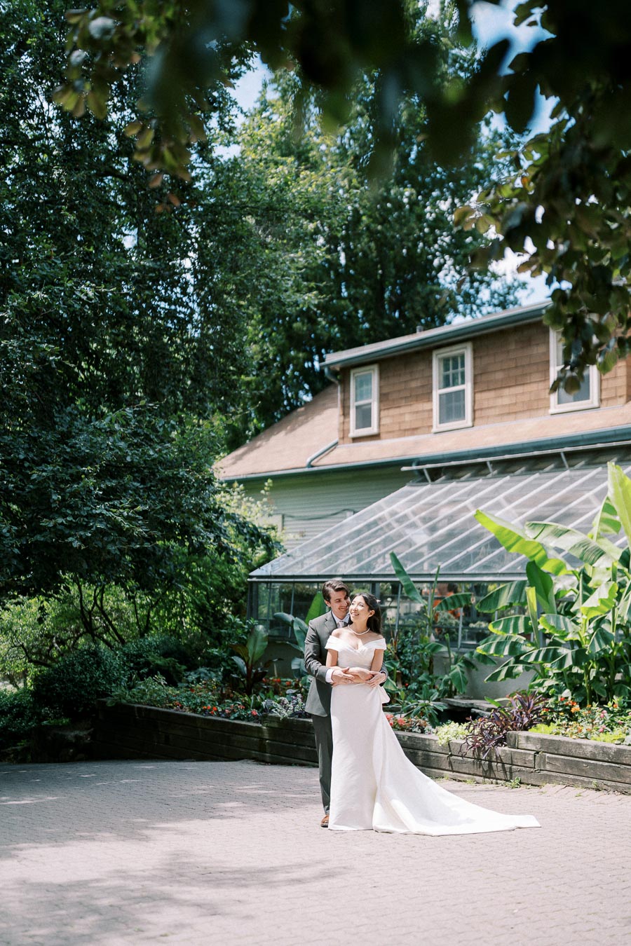 A bride and groom embrace in a lush garden setting, standing on a paved pathway near a quaint wooden greenhouse, surrounded by vibrant green foliage and colorful flowers, on a sunny day.