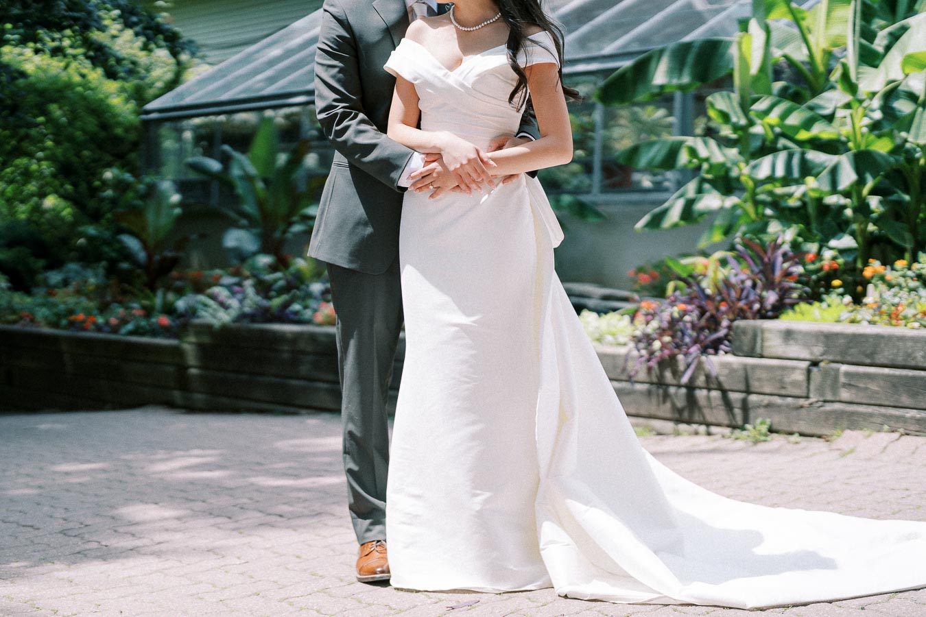 Wedding couple posing outdoors in elegant attire, with the bride wearing a white gown and the groom in a dark suit, surrounded by lush greenery and colorful flowers.