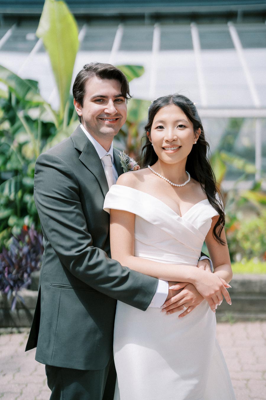 Bride and groom smiling at their wedding, with greenery and greenhouse background, she in an off-shoulder white dress and pearl necklace, he in a dark suit and tie.