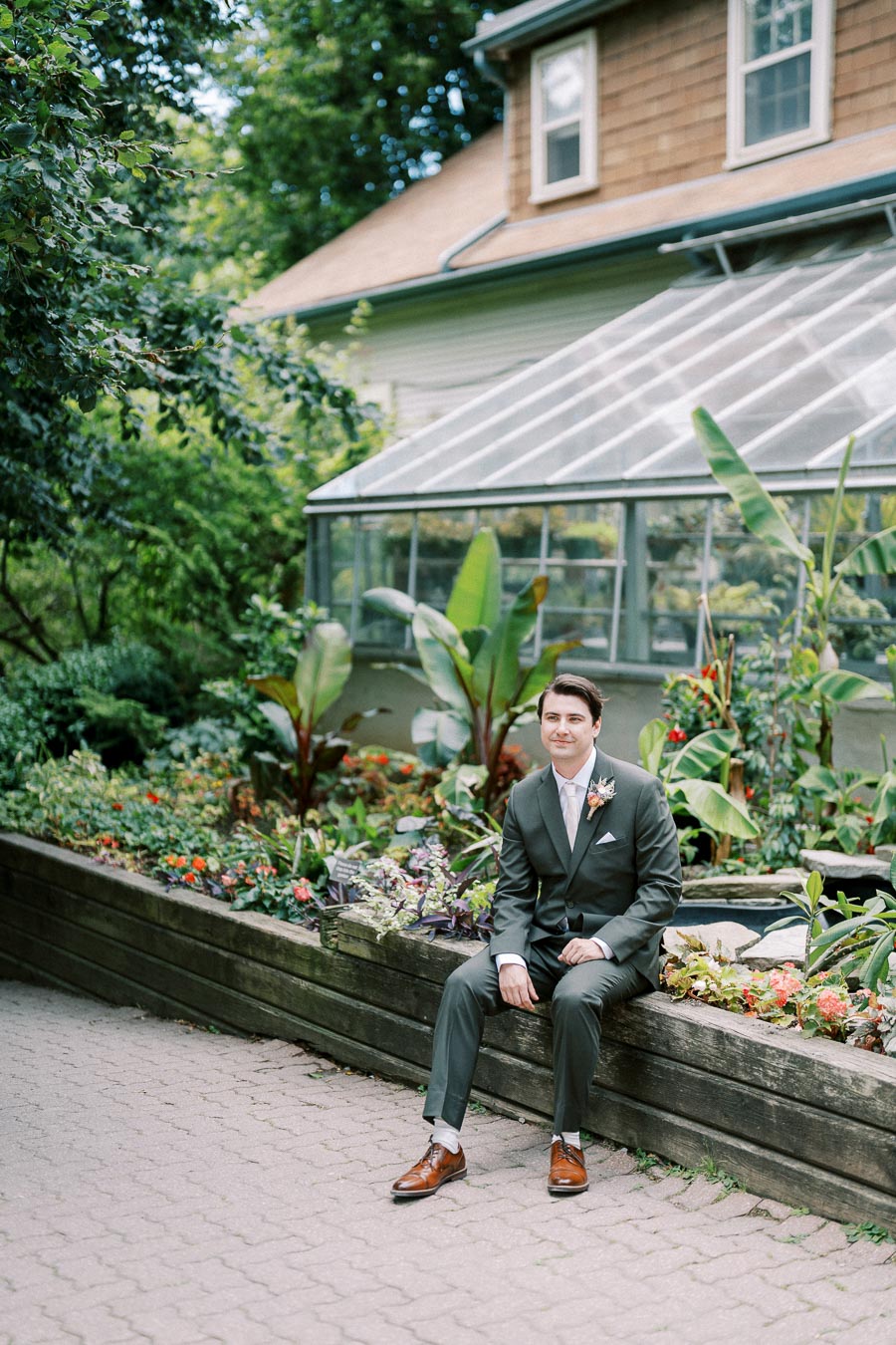 Man in a suit sitting on a garden ledge with lush green plants and a greenhouse in the background.