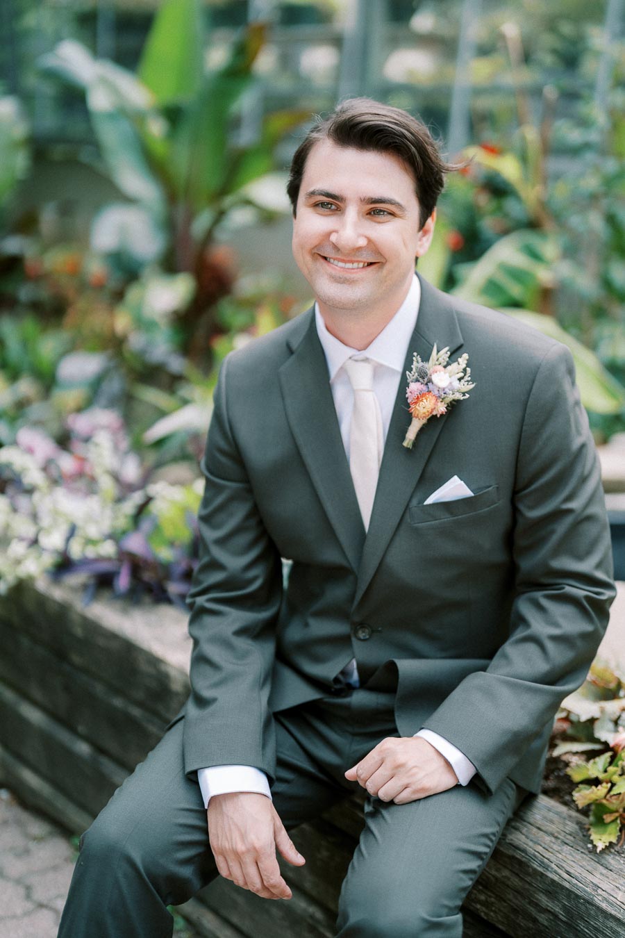 Smiling groom in a dark suit with a boutonniere, sitting on a wooden bench surrounded by lush greenery and colorful flowers, exuding joy and elegance.
