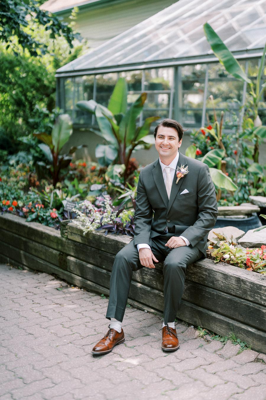 Man in gray suit sitting on a wooden ledge in a garden setting with greenhouse in background, smiling and surrounded by lush green plants and flowers