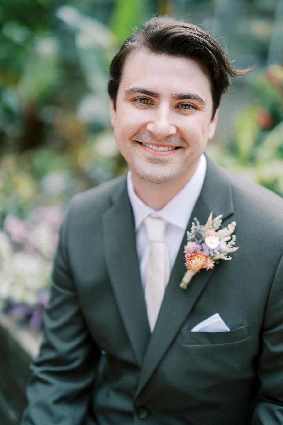 A smiling man in a dark green suit with a floral boutonnière, sitting outdoors in a lush garden setting, exuding joy and elegance.
