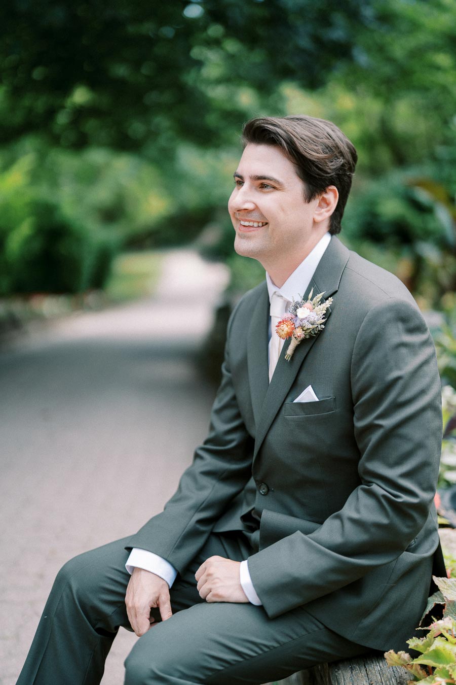Smiling man in a dark suit sitting on a bench in a lush garden setting, wearing a boutonniere with pink and orange flowers, showcasing a joyful expression.