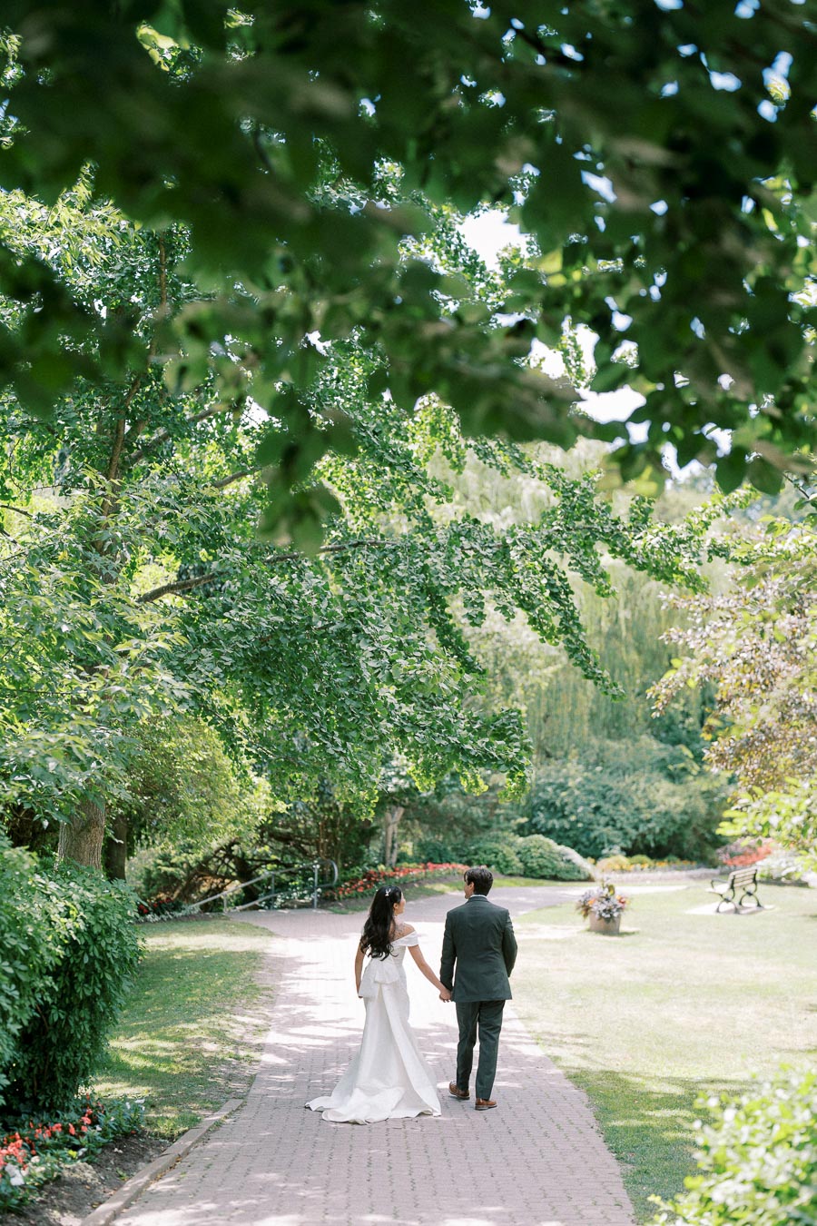 A bride and groom walking hand in hand down a garden path, surrounded by lush green trees and colorful flowers, on a sunny day.
