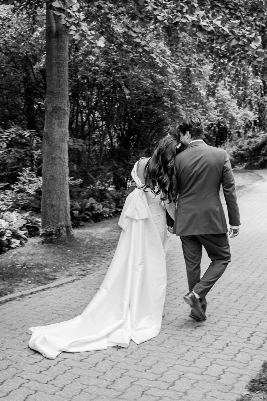 A newlywed couple walking arm in arm down a tree-lined path, with the bride in a flowing white gown and the groom in a dark suit, captured in a romantic black and white photograph.