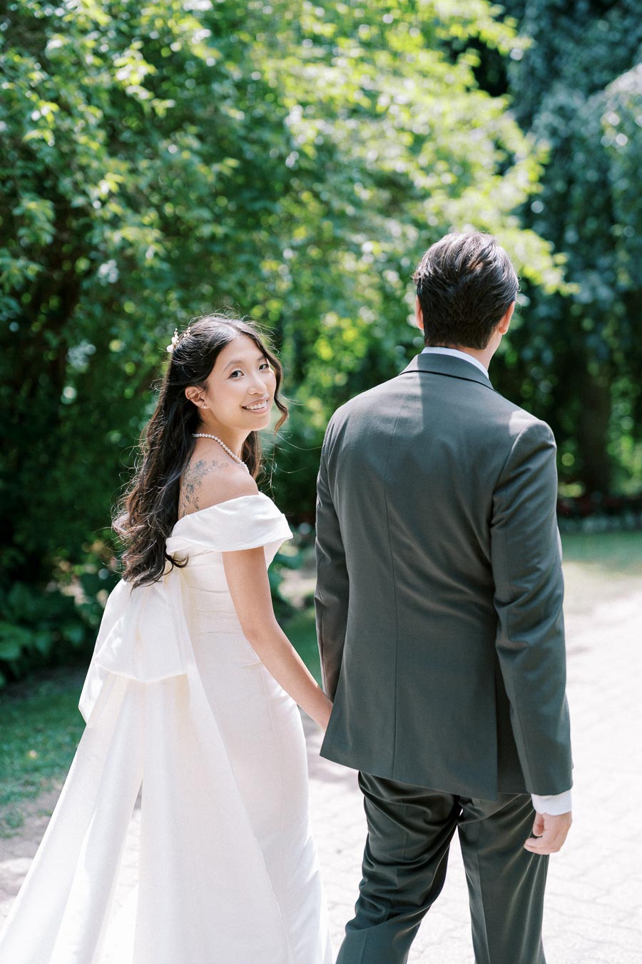 Smiling bride in elegant white gown walking hand in hand with groom in dark suit along a sunlit garden path, surrounded by lush greenery.