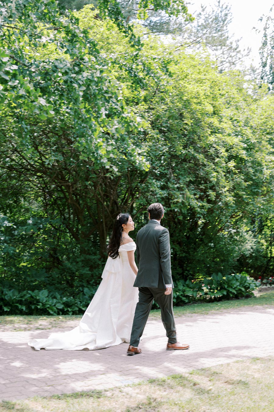 A bride and groom walking together on a garden path surrounded by lush greenery and sunlight, capturing a romantic wedding moment.