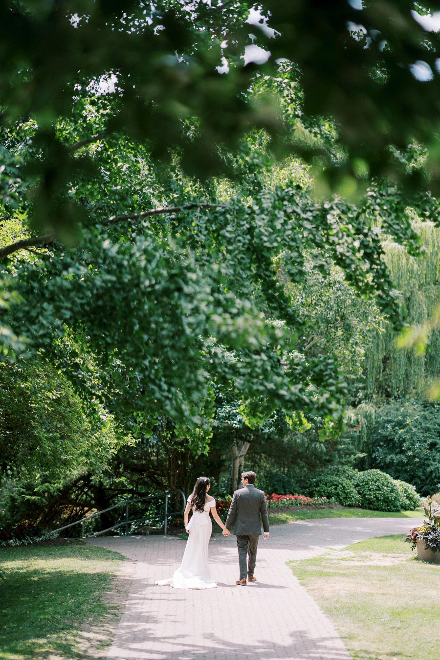 Couple walking hand in hand under lush green trees on a sunny day, enjoying a romantic stroll in a garden setting.