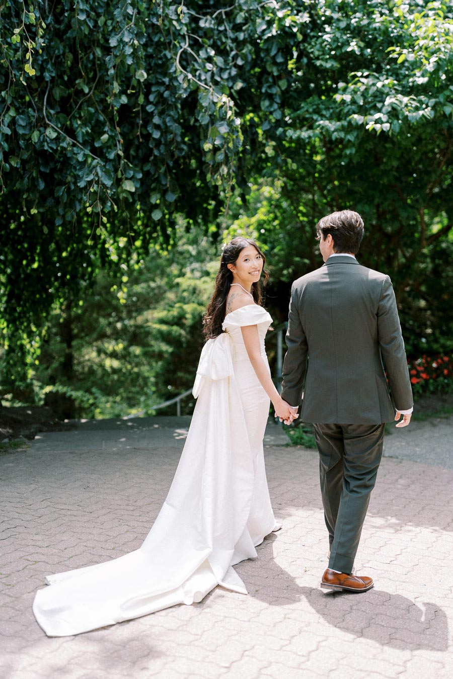A bride in an elegant white wedding gown with a long train holds hands with her groom in a dark suit as they walk down a tree-lined pathway, surrounded by lush greenery and sunlight.