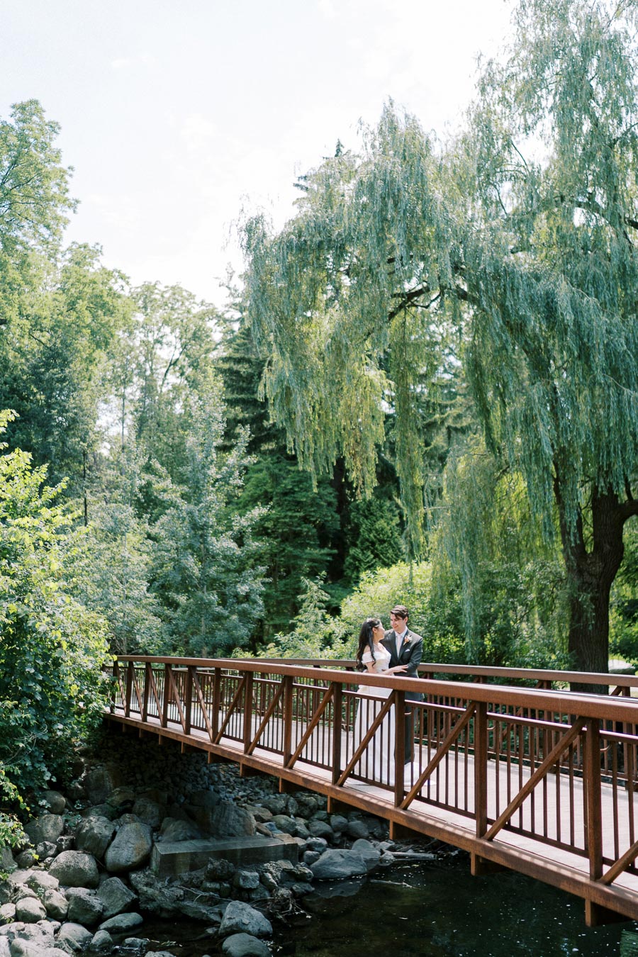 A couple in formal attire stands on a wooden bridge surrounded by lush greenery and tall trees, creating a serene and romantic atmosphere.