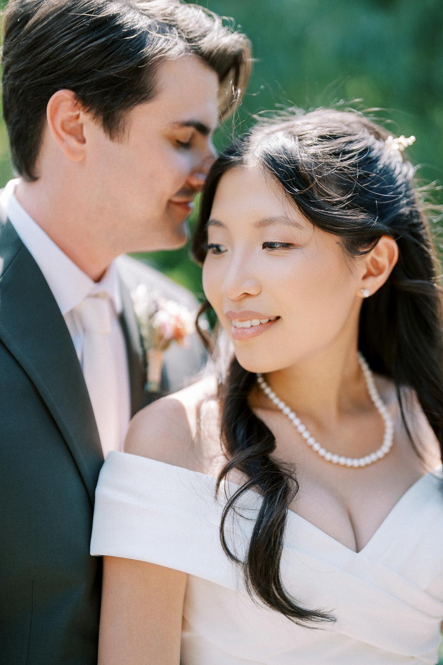 Romantic wedding portrait of a couple in elegant attire, with the groom in a gray suit and the bride wearing a white off-shoulder dress and pearl necklace, set against a lush green background.