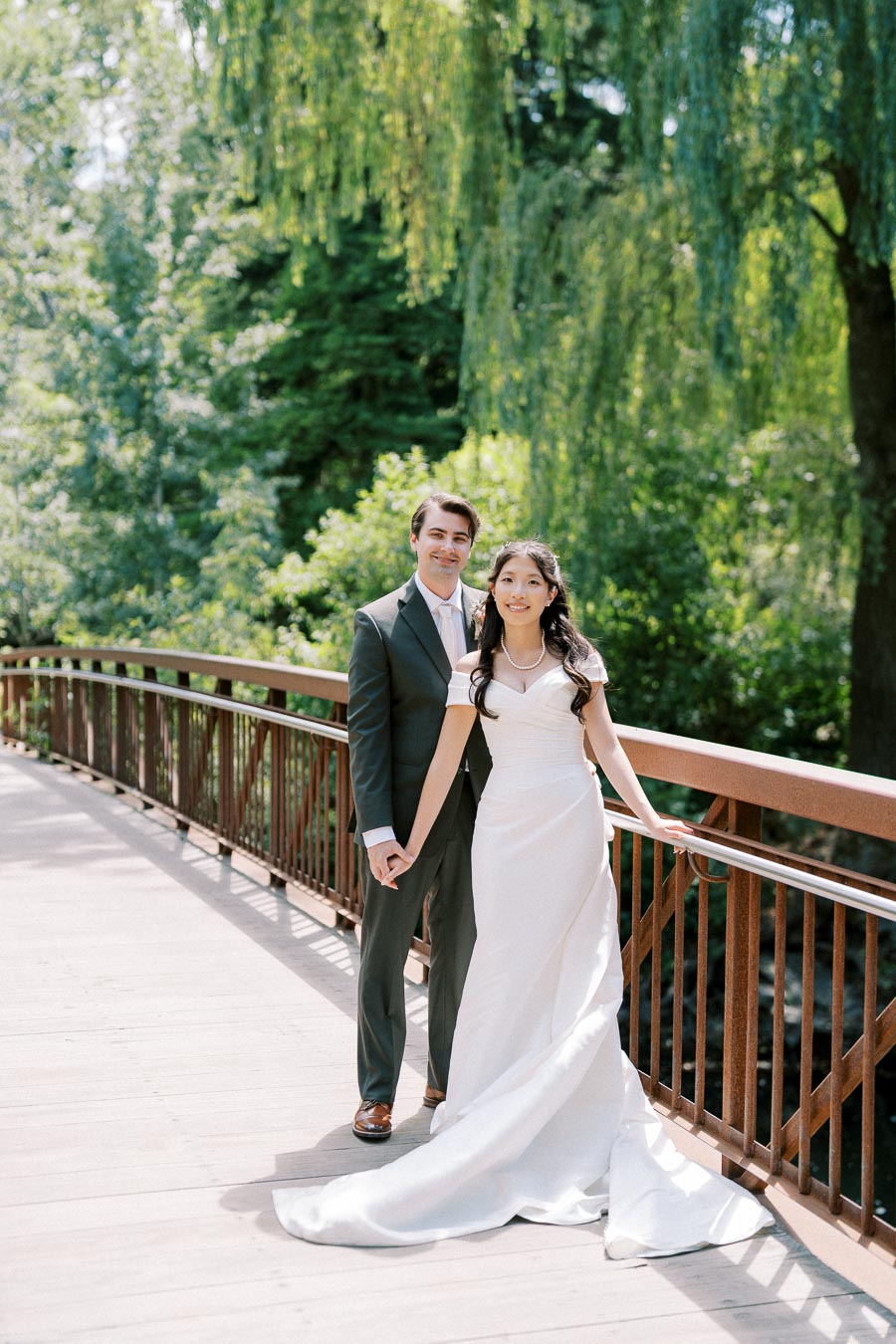 A bride and groom standing on a rustic wooden bridge surrounded by lush greenery, captured during their outdoor wedding photoshoot.