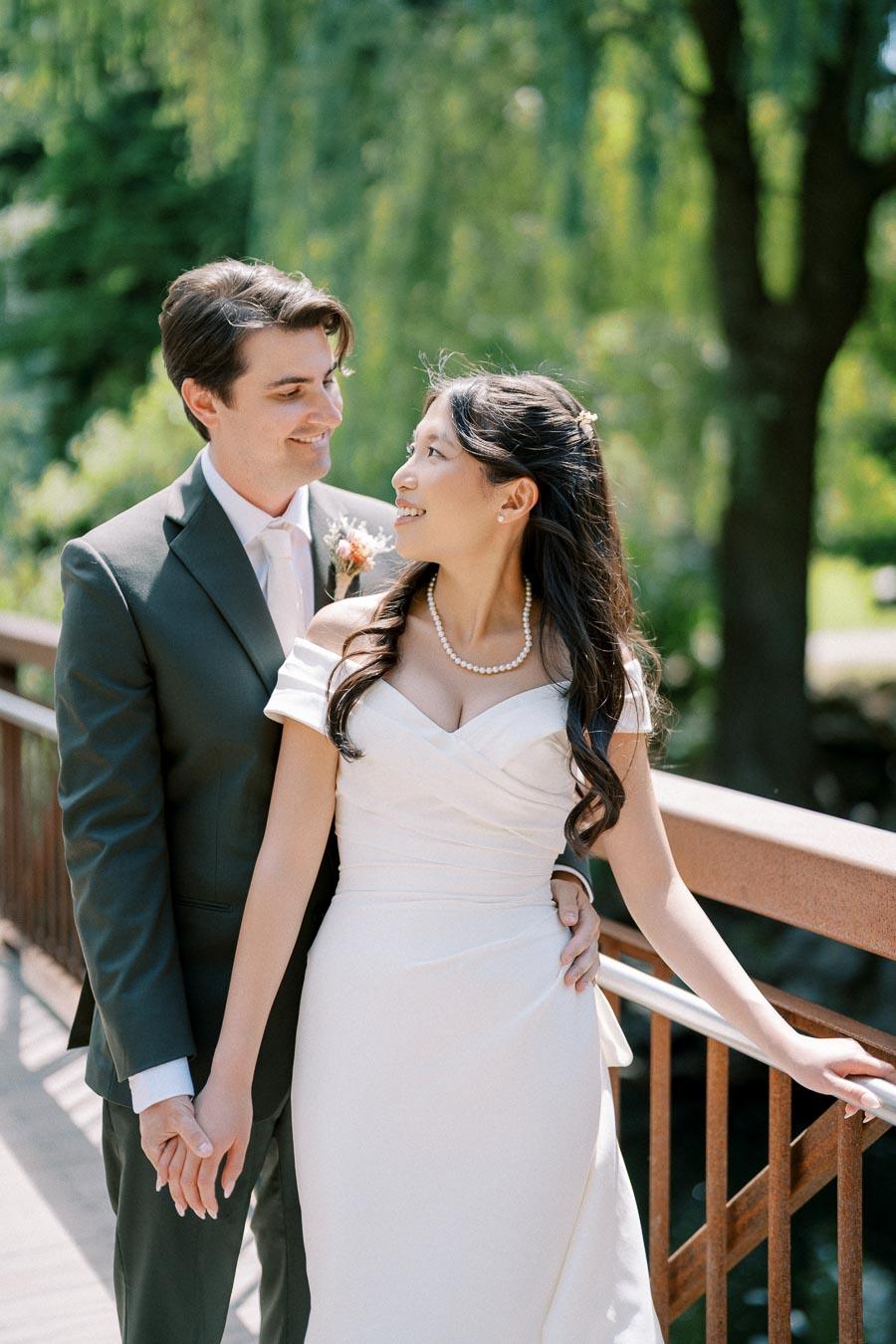 A bride and groom standing on a bridge in a picturesque outdoor setting, holding hands and looking at each other lovingly, with lush green trees in the background.