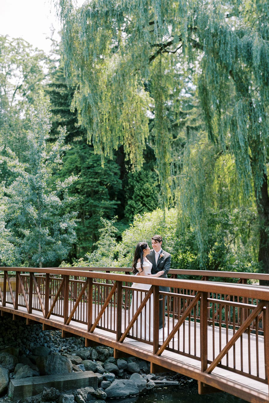 A couple embraces on a wooden bridge surrounded by lush green trees in a scenic outdoor park setting.