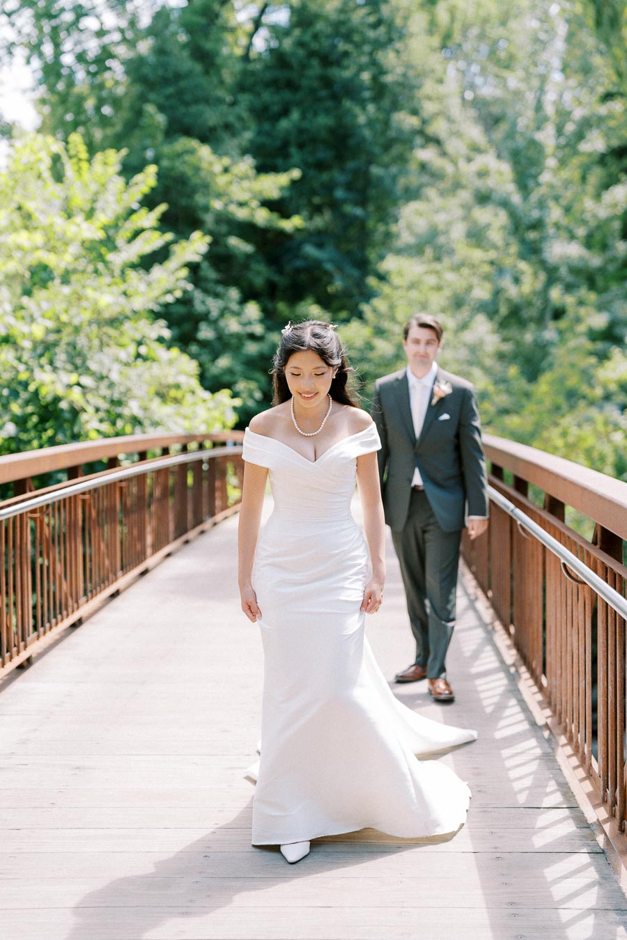 Bride and groom on wooden bridge in lush garden setting, with bride in elegant white gown leading and groom in suit following, capturing a romantic wedding moment.