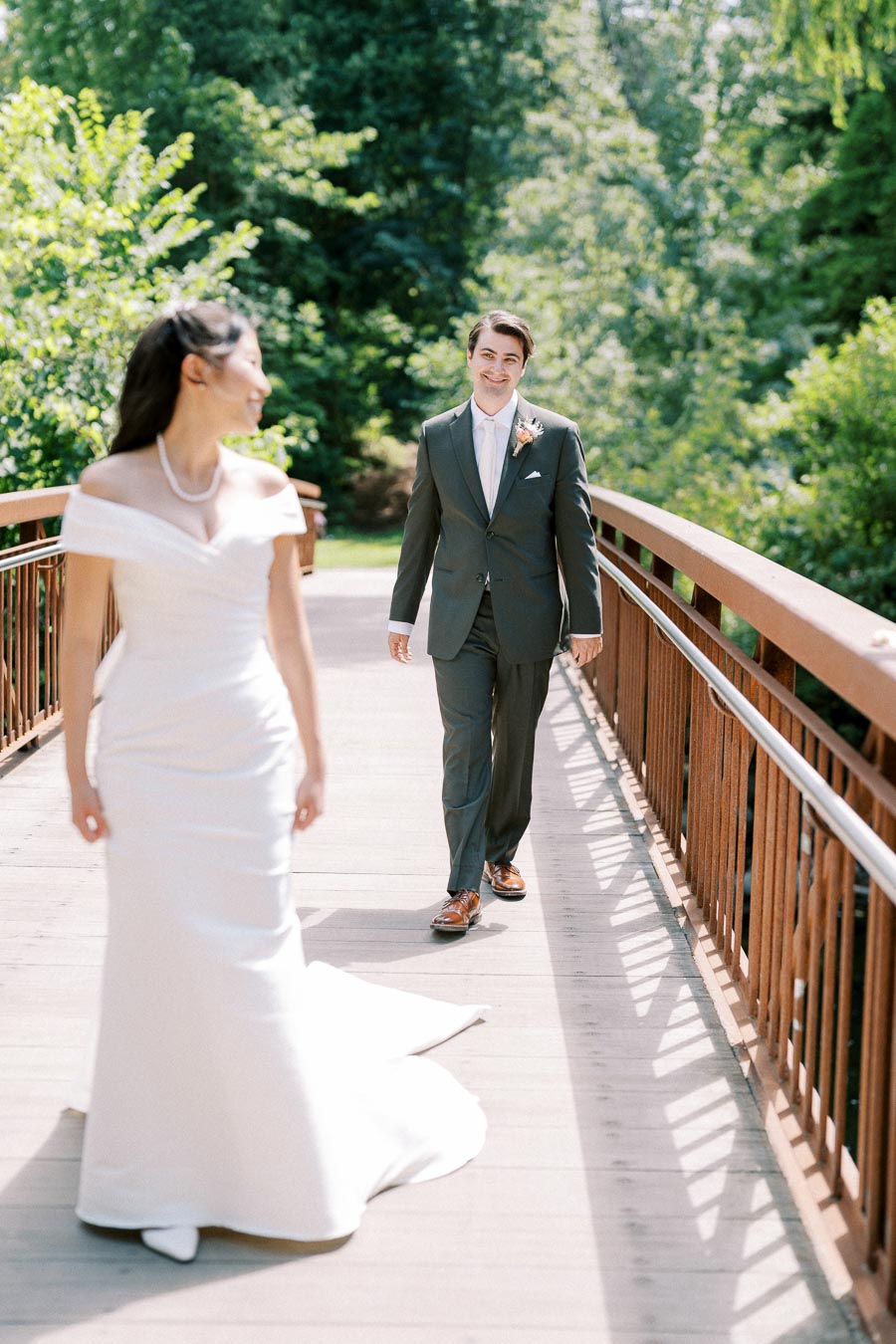 A bride in a white dress smiles as she walks ahead of a groom in a dark suit on a wooden bridge surrounded by lush greenery.