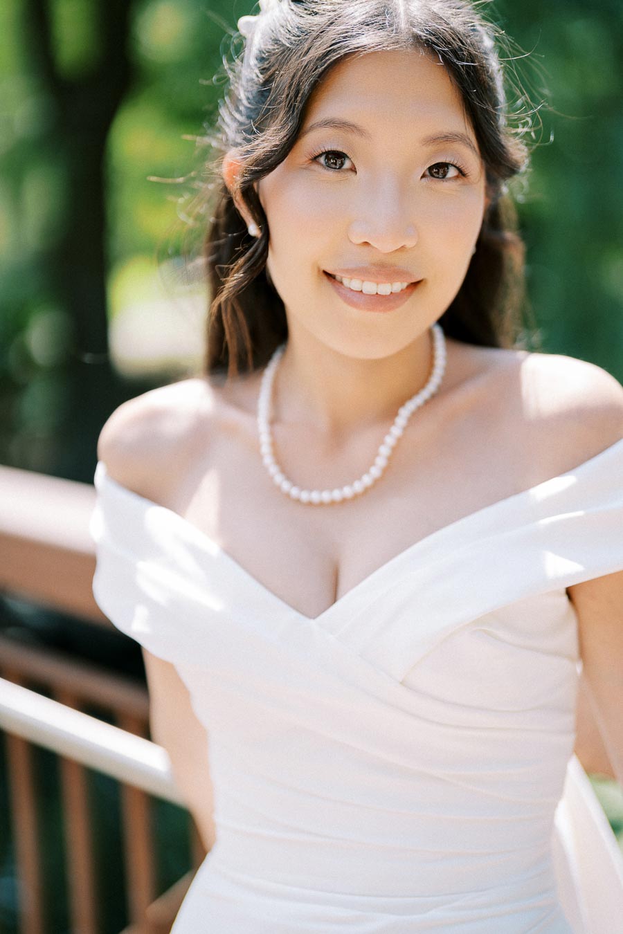A smiling bride in an elegant off-shoulder white gown and pearl necklace, standing outdoors with a blurred green background.