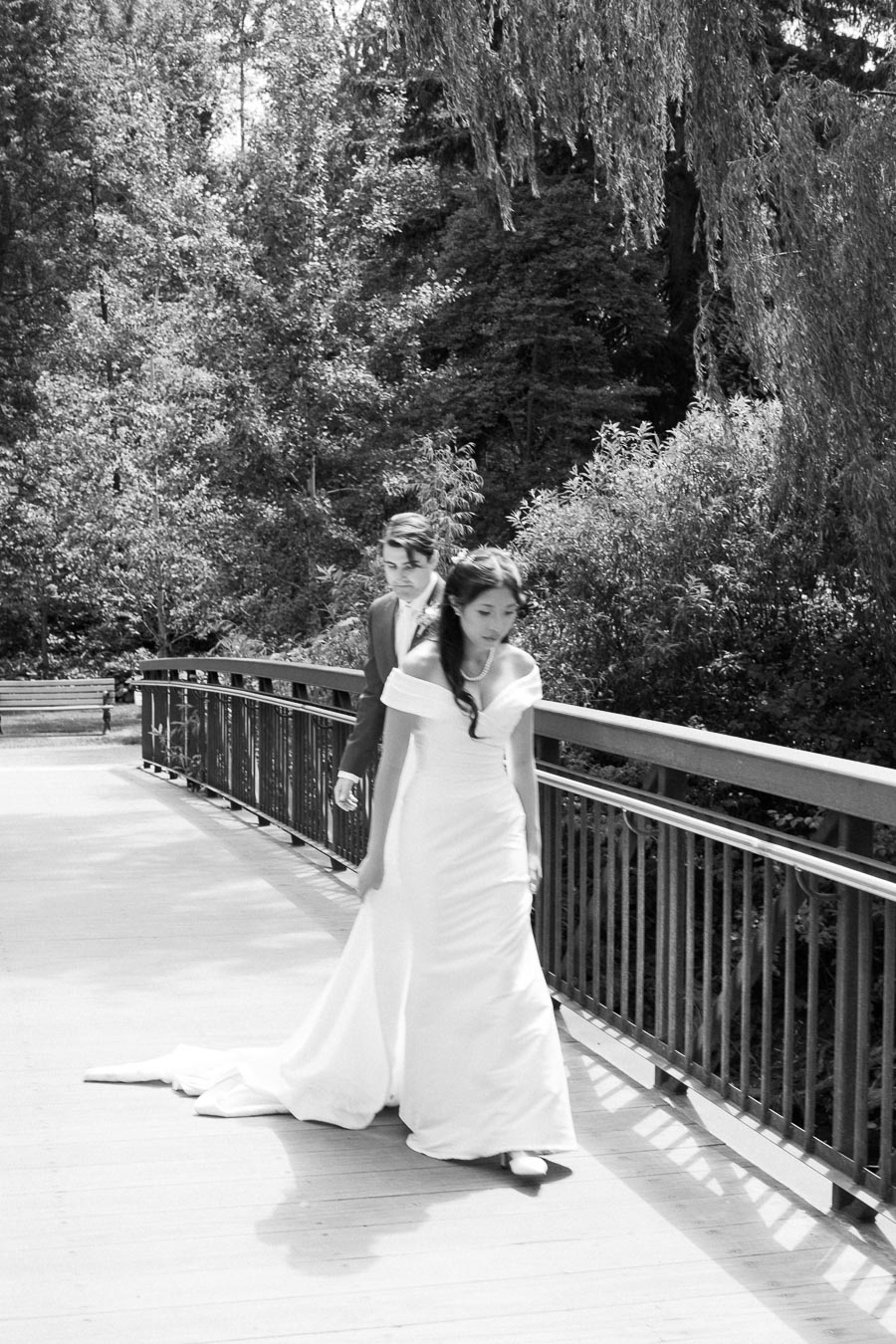 Black and white photo of a couple walking on a bridge in a lush, wooded park setting. The woman is wearing an elegant white wedding gown and the man is in a suit, capturing a serene, romantic moment.