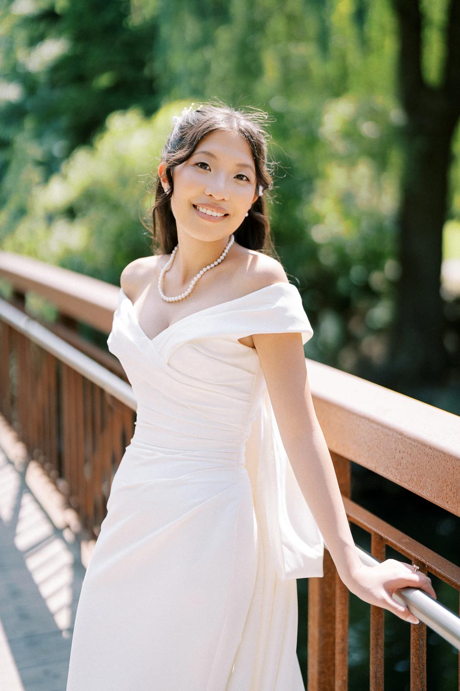 Smiling bride in an elegant white wedding dress standing on a bridge surrounded by lush greenery