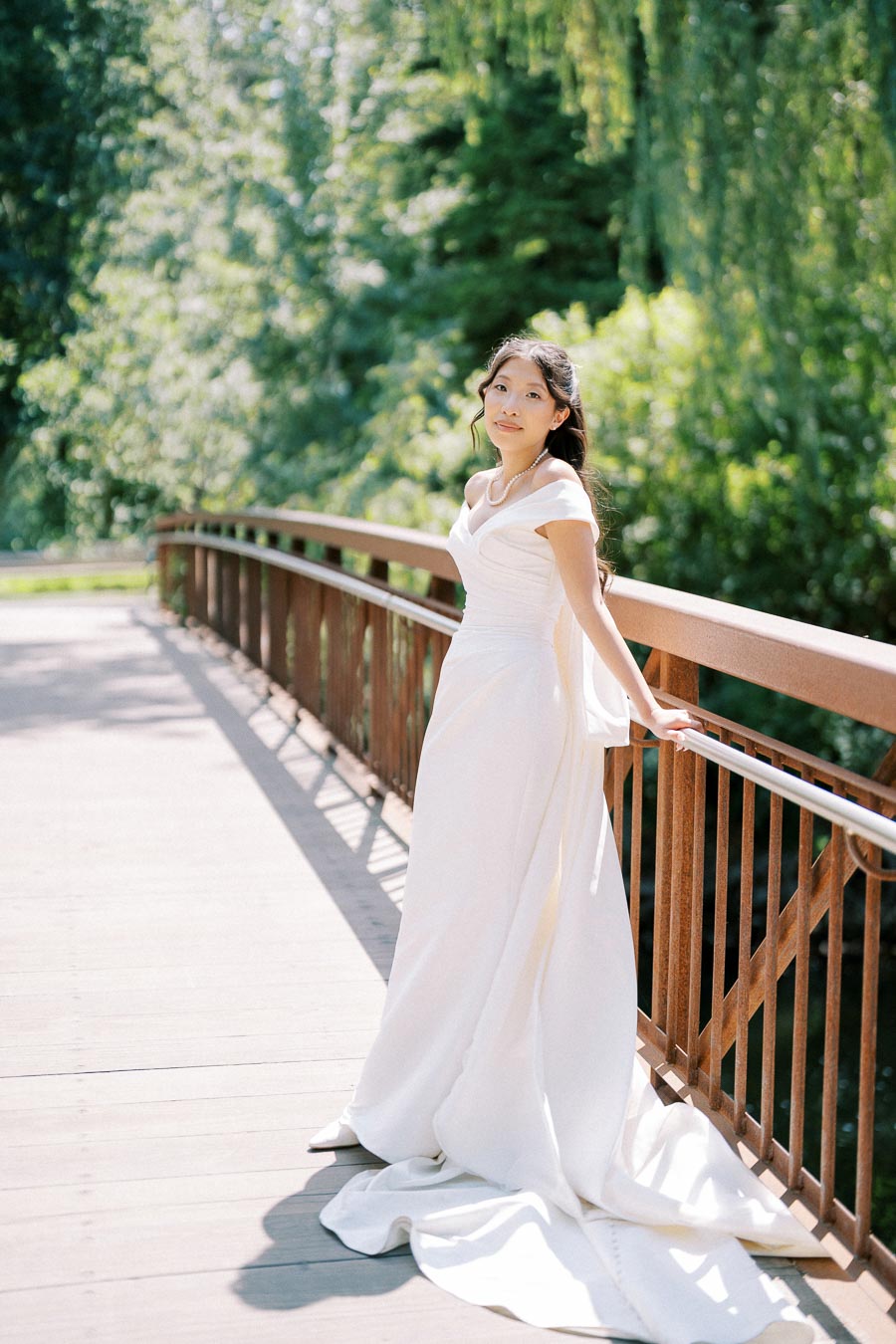 A bride in a flowing white gown stands gracefully on a wooden bridge, surrounded by lush greenery and dappled sunlight, capturing a serene and romantic moment in nature.
