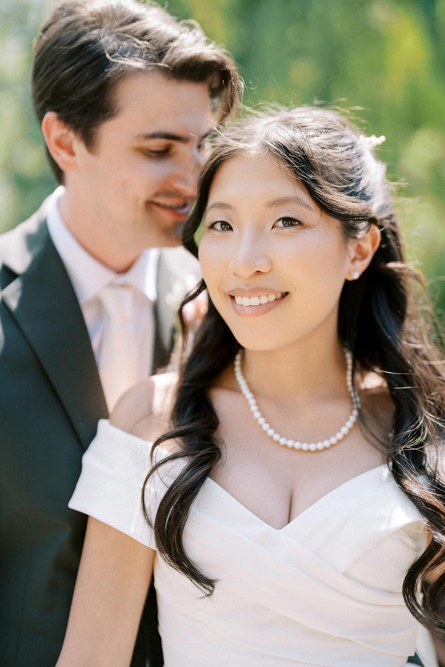 Smiling bride in white dress with pearl necklace and groom in suit embracing outdoors