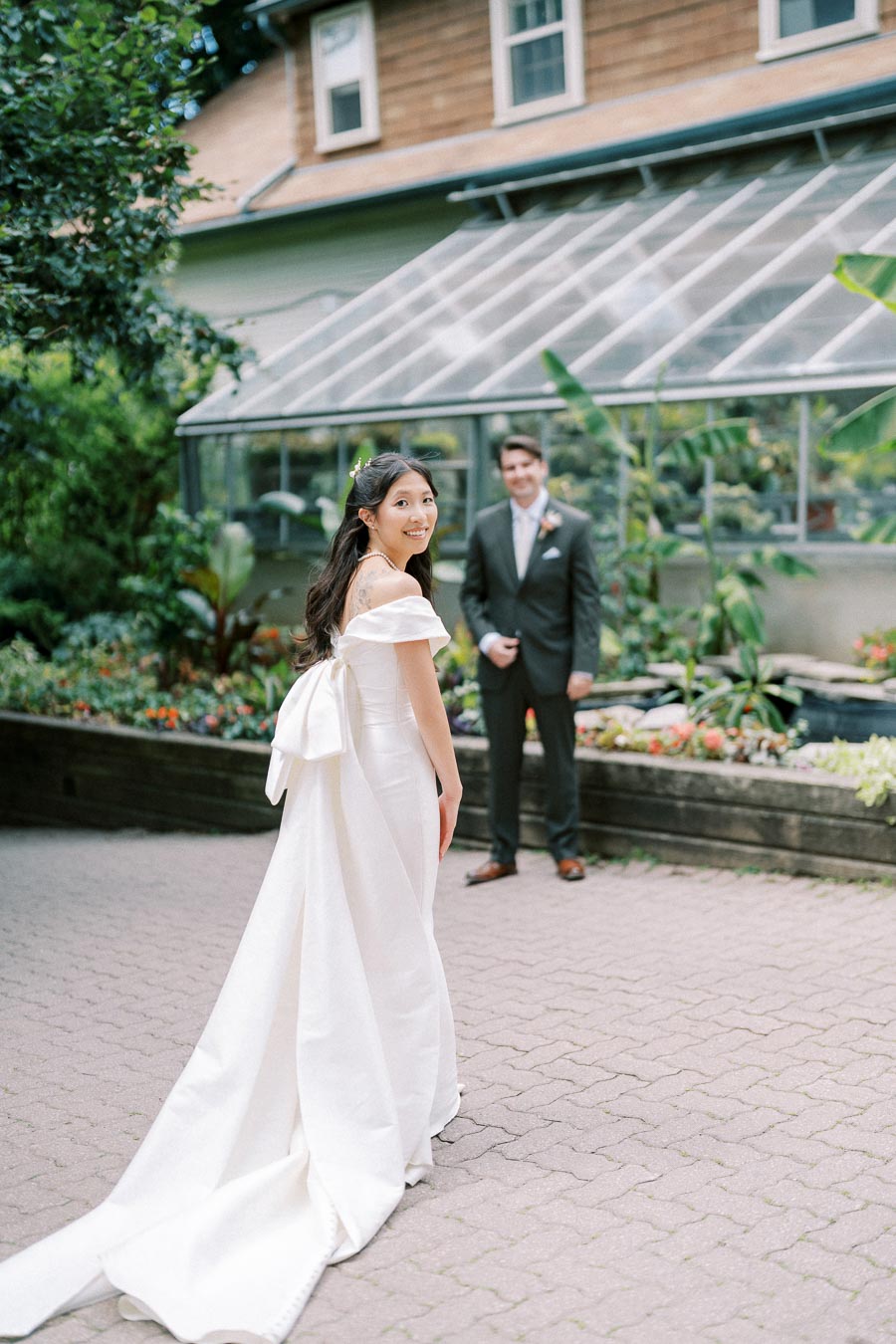 Bride in elegant white wedding dress with long train smiles as she looks back at the groom standing in front of a greenhouse, surrounded by lush greenery and vibrant flowers.