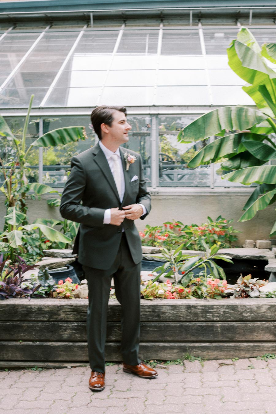 A groom in a dark suit stands in a greenhouse surrounded by vibrant green plants and tropical foliage.