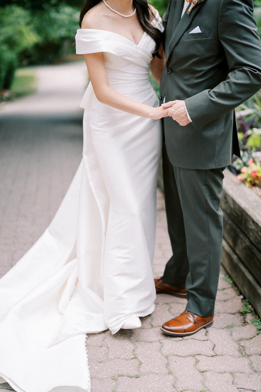 Wedding couple holding hands, bride in an elegant off-the-shoulder white gown and groom in a dark suit, standing on a garden pathway.