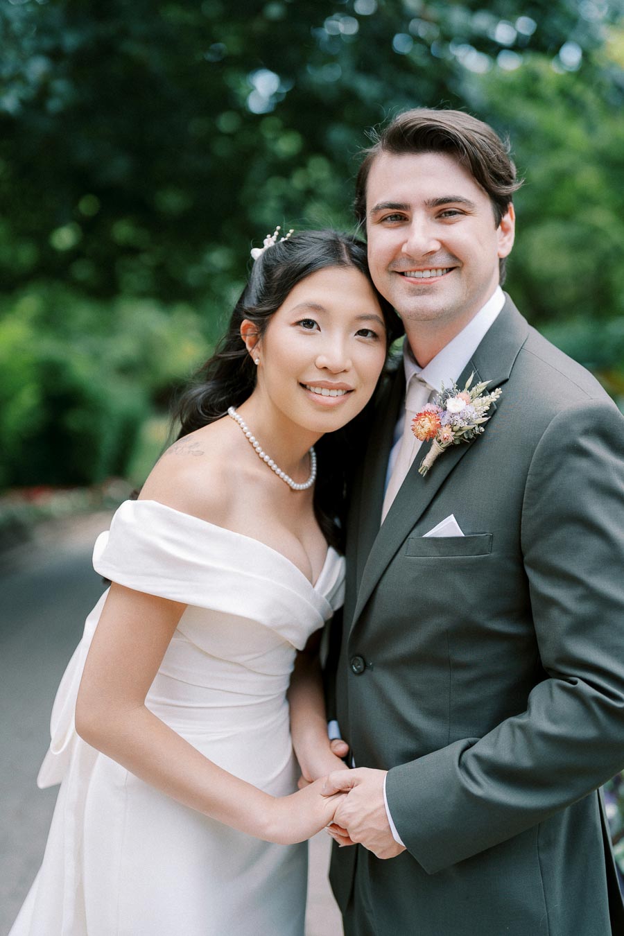 A happy couple in wedding attire posing outdoors; the bride is wearing an elegant white dress with pearl necklace, and the groom is in a dark suit with a boutonniere, standing together in a lush green garden.