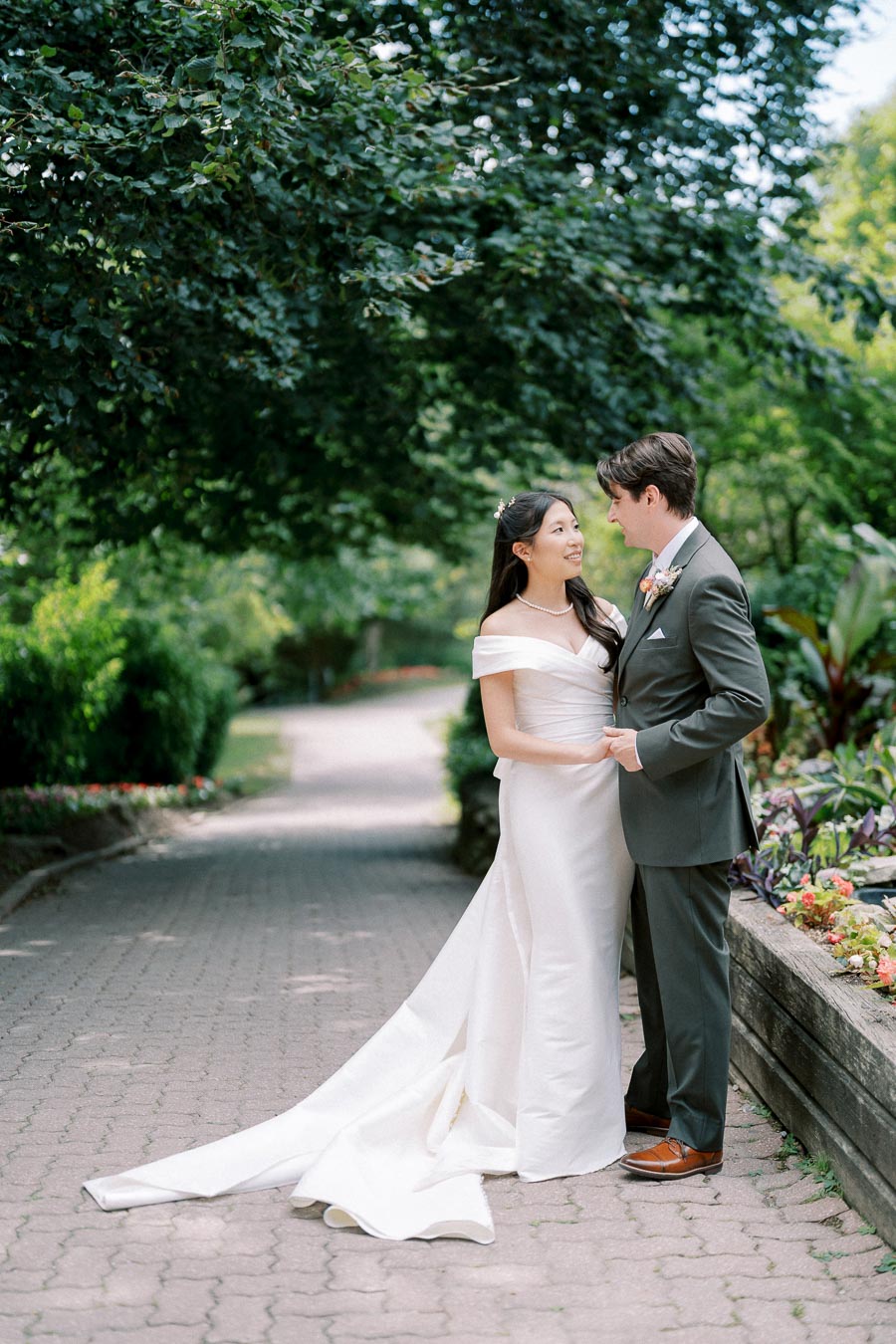 A bride in an elegant white wedding dress and a groom in a dark suit share a moment in a beautiful garden setting.