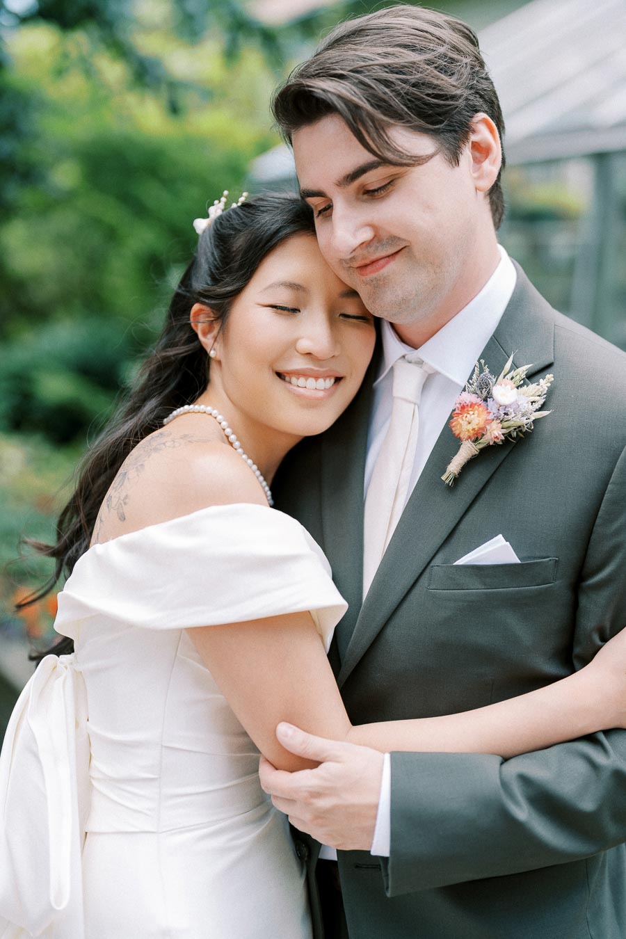 A joyful bride and groom embracing on their wedding day, surrounded by lush greenery, with the bride wearing an elegant white gown and pearl necklace, and the groom in a classic dark suit with a floral boutonniere.