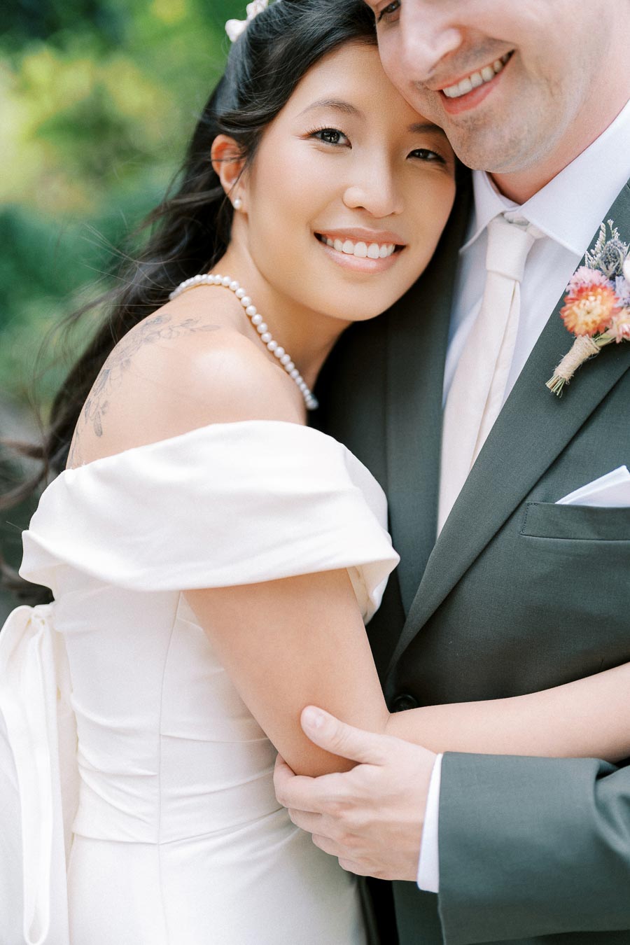 A bride in an elegant white dress smiles while embracing her groom, who is wearing a dark suit with a floral boutonniere. The couple appears joyful and radiant on their wedding day, surrounded by a green, natural backdrop.