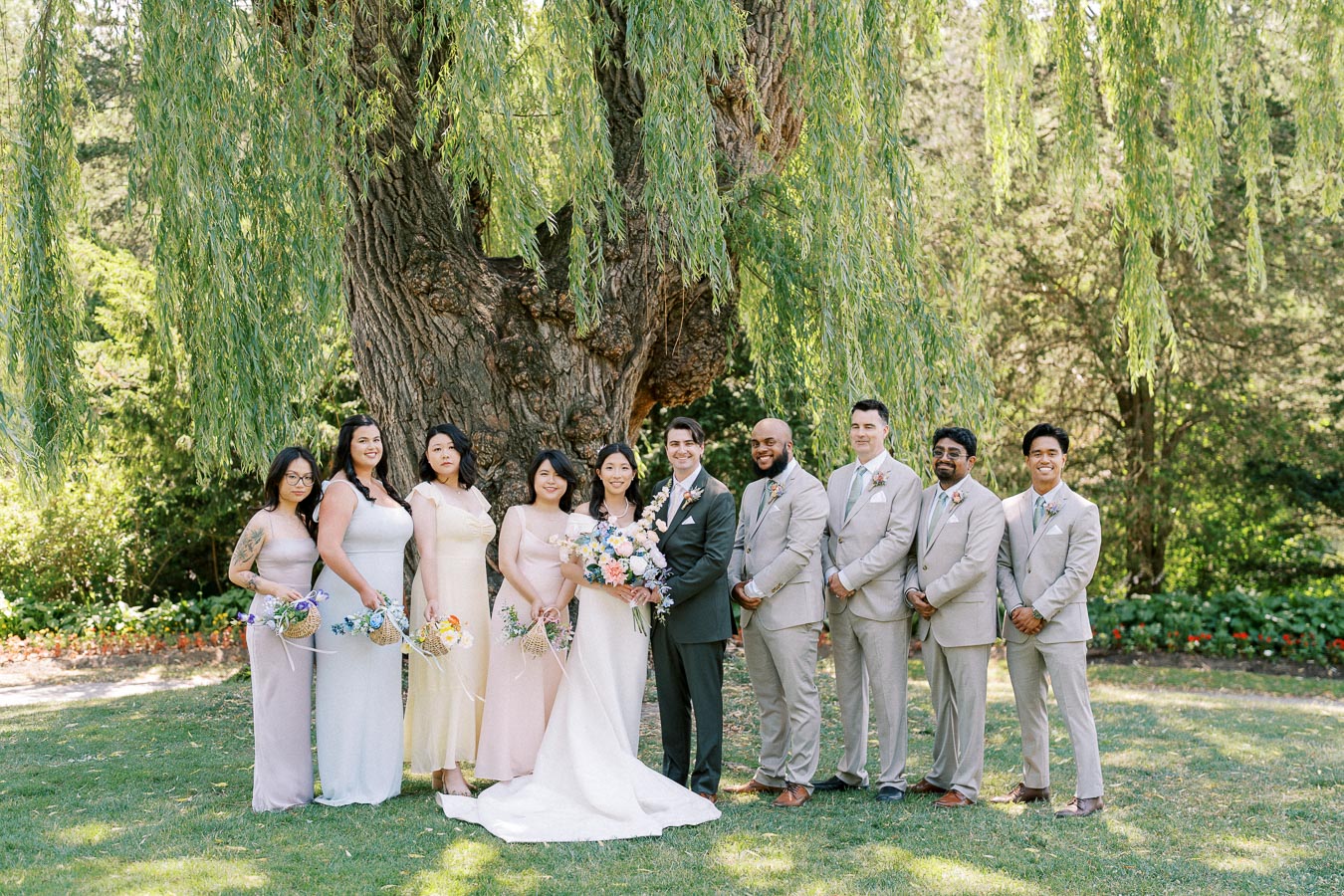 Wedding party posing under a large tree, with the bride in a white gown holding a colorful bouquet, surrounded by bridesmaids in pastel dresses and groomsmen in light grey suits, in a lush garden setting.