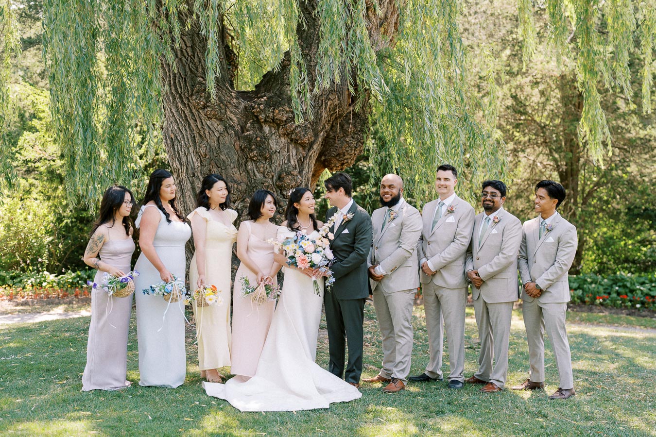 Wedding party posing under a large willow tree, featuring bridesmaids in pastel dresses and groomsmen in beige suits, celebrating a sunny outdoor ceremony.