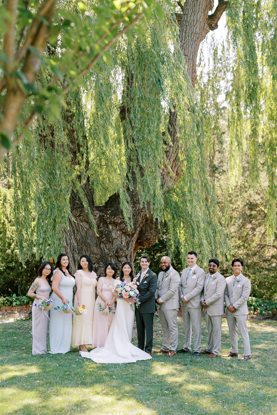 Wedding party posing outdoors under a large tree, featuring the bride in a white gown and groom in a dark suit, surrounded by bridesmaids in pastel dresses and groomsmen in light suits, all holding bouquets.