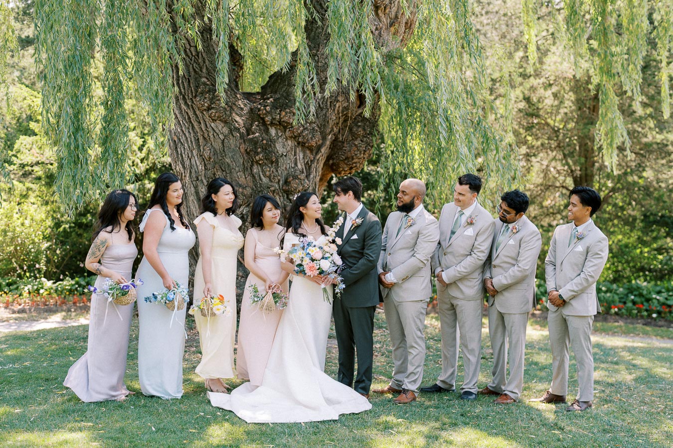Wedding party posing outdoors under a large tree, featuring the bride in a white gown holding a bouquet of flowers, bridesmaids in pastel dresses, and groomsmen in light gray suits, all creating a harmonious and joyful scene.