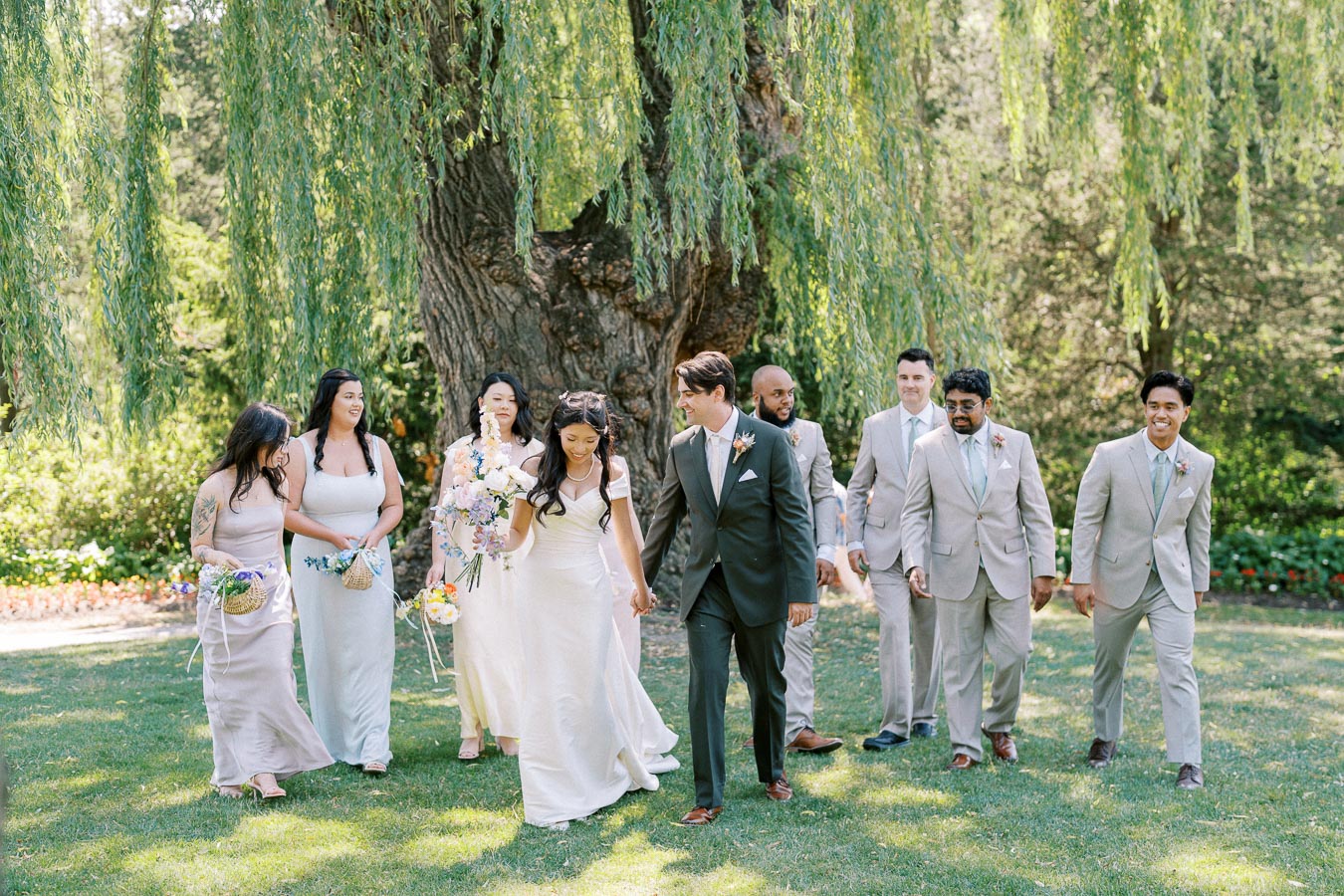 A joyful wedding party celebrating under a lush willow tree in a sunny garden. The bride, wearing a white gown, walks hand in hand with the groom in a dark suit, surrounded by bridesmaids in pastel dresses and groomsmen in light-colored suits, capturing the essence of a beautiful outdoor wedding ceremony.
