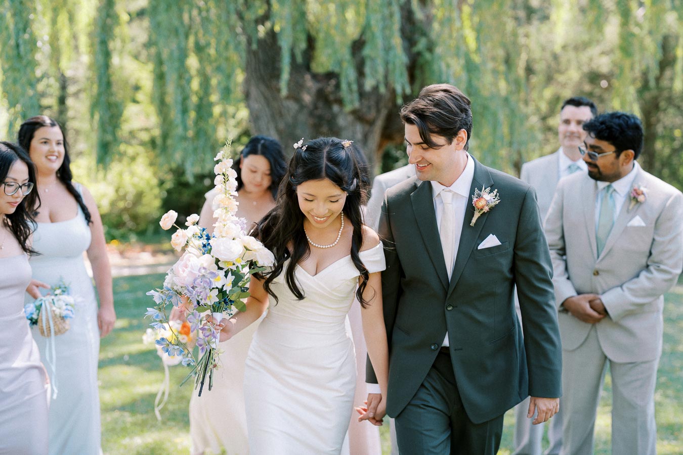 Wedding couple holding hands outdoors, surrounded by bridal party in elegant attire, under lush greenery, with bride holding a pastel floral bouquet.