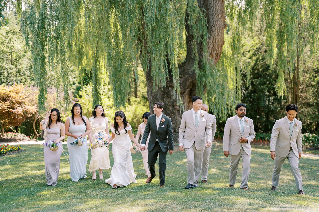 A wedding party walks joyfully together in a lush garden under a large willow tree, with bridesmaids wearing pastel dresses and groomsmen in light suits.