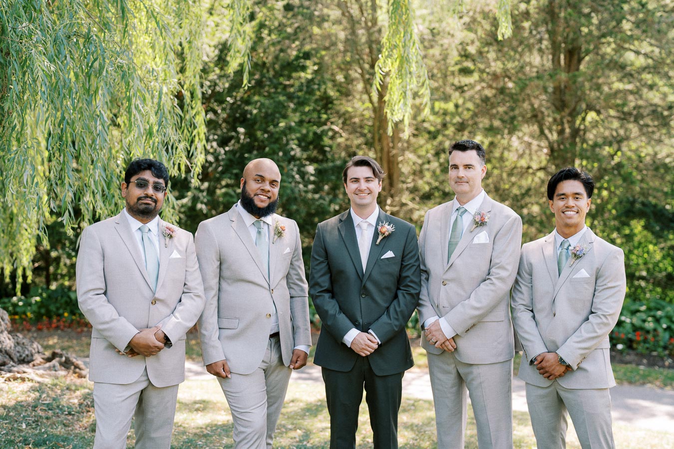 Five men dressed in formal suits posing outdoors in a lush green garden setting, ideal for a wedding or special event showcasing groomsmen fashion.