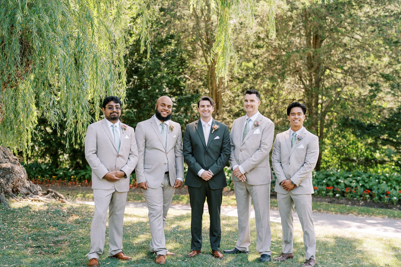 Five groomsmen in light gray suits and a groom in a dark green suit stand smiling under a lush willow tree in a garden setting.