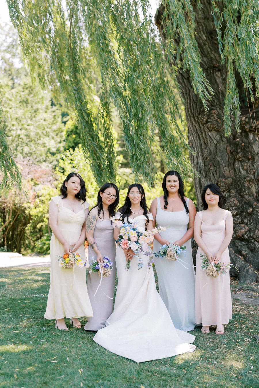 A group of five women in pastel dresses standing together outdoors under a willow tree, each holding a bouquet, with the bride in the center wearing a white wedding gown and a serene garden setting in the background.