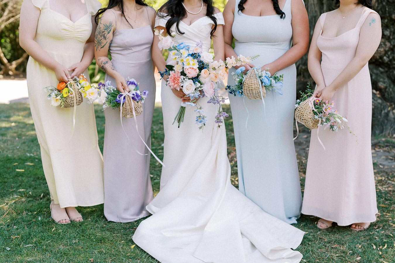Bridesmaids wearing pastel dresses holding floral bouquets while standing on grass at an outdoor wedding.
